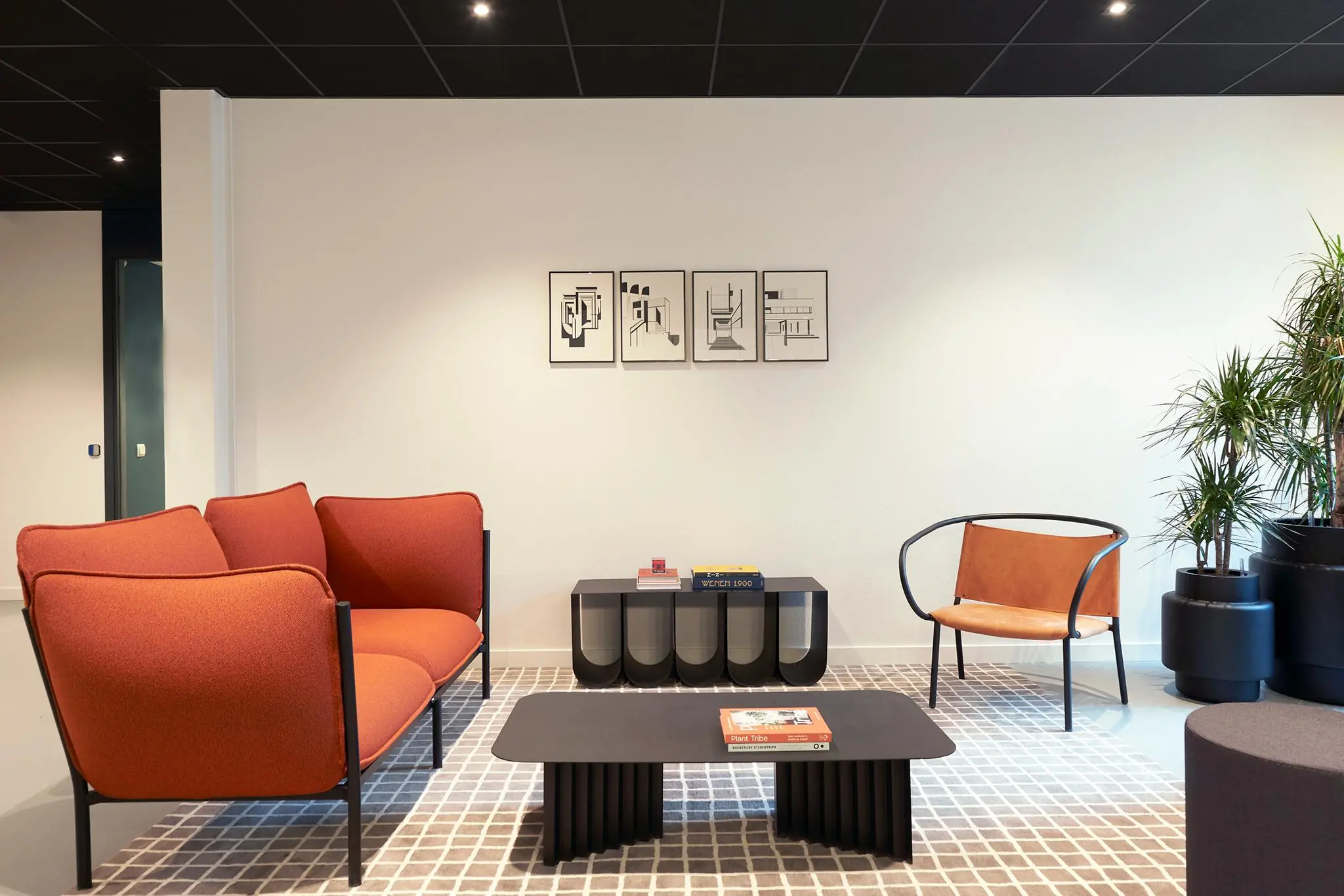 Modern ingerichte wachtruimte met een oranje bank, designstoel, zwarte salontafel, kunst aan de muur en grote kamerplanten.